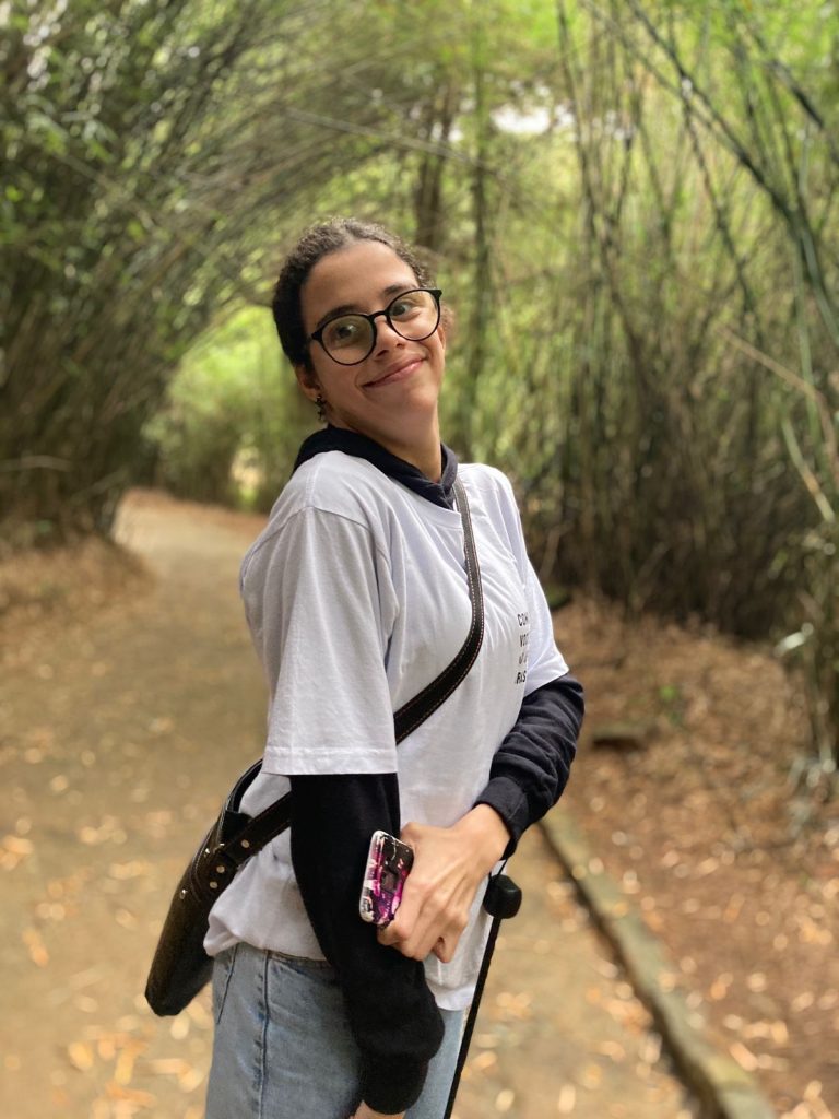 A jovem Ana Laura está de camiseta branca, uma blusa por baixo de mangas longas e uma bolsa preta de lado. Ela é branca, tem cabelos escuros e está usando óculos de grau pretos redondos. 